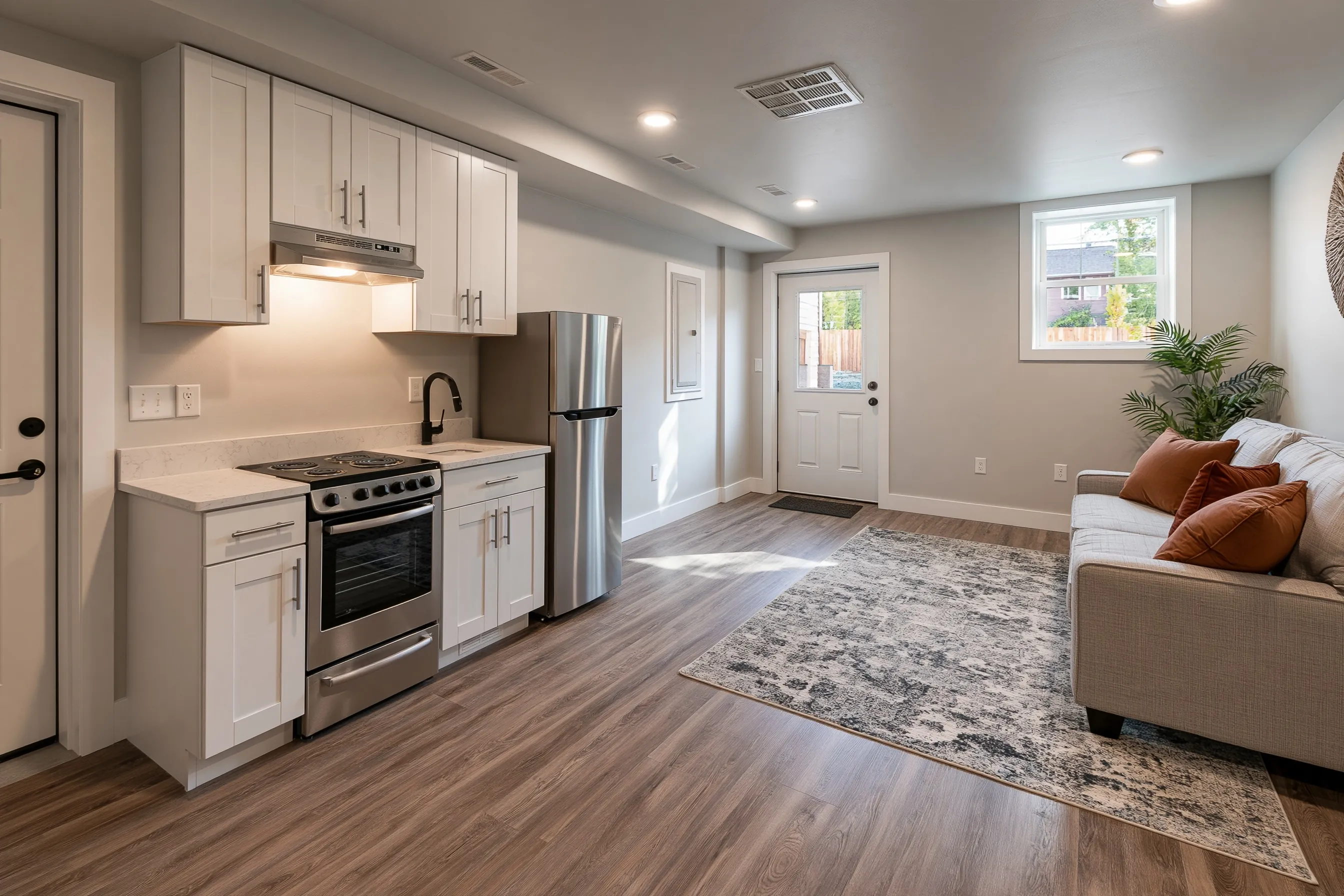 Basement apartment main room with a kitchenette along one wall, an apartment-size refrigerator, a sofa and a separate exterior door with a glass panel on the far side